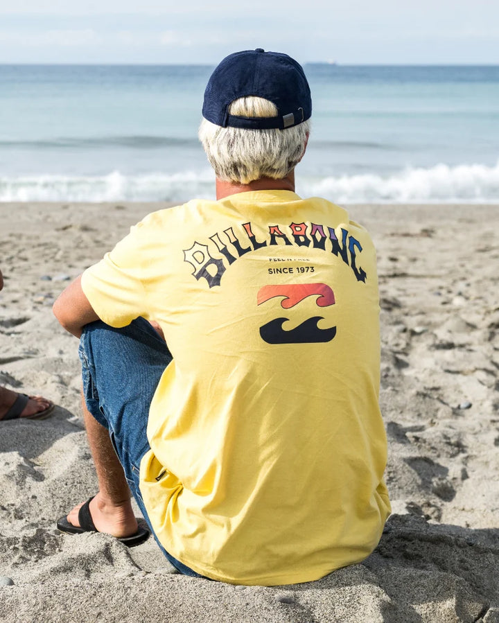 Man sitting on a beach wearing a yellow Billabong t-shirt.